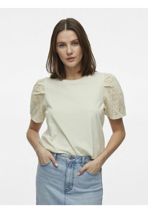 Camiseta de color beige claro con mangas cortas abullonadas. Las mangas tienen detalles de ojal. Tejido suave, diseño de cuello redondo, combinada con una falda de mezclilla.