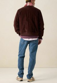 Bruine corduroy jas met een ribbelstructuur, gecombineerd met lichtblauwe spijkerbroeken en beige sneakers. Opvallend geruite kraagdetail.