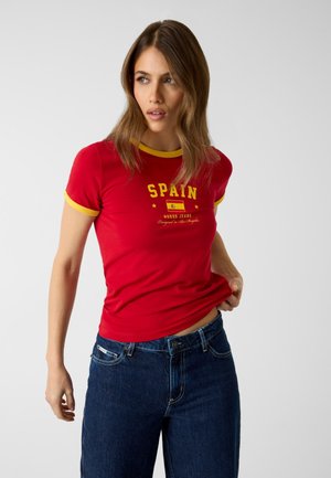 Kobieta ubrana w czerwony dopasowany T-shirt z żółtym wykończeniem i napisem „Hiszpania”, w połączeniu z niebieskimi jeansami, pozująca na jednolitym tle.