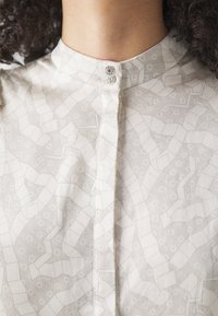 Blusa leggera color crema con motivo geometrico testurizzato. Presenta un colletto rotondo e due bottoni decorativi al collo.
