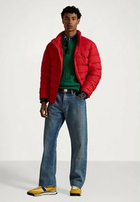 Muškarac u crvenoj puffer jakni, zelenom džemperu, plavim farmerkama i žutim sportskim cipelama stoji s jednom rukom u džepu.