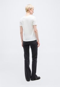 Άτομο με κοντά ξανθά μαλλιά στέκεται γυρισμένο προς τα πίσω, φορώντας άσπρο κοντομάνικο πουκάμισο, μαύρα τζιν και μαύρα παπούτσια σε απλό φόντο.