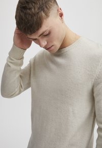 Joven con cabello corto y castaño, vistiendo un suéter de punto crema, tocándose la cabeza y mirando hacia abajo contra un fondo claro y liso.