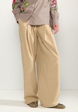Personne portant un pantalon large beige ample, des chaussures marron décontractées, et une chemise rayée avec une broderie florale sur les manches.
