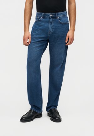 Mężczyzna w prostych, niebieskich dżinsach, czarnych mokasynach i czarnej koszuli wpuszczonej do spodni, stojący na jasnym, jednolitym tle.