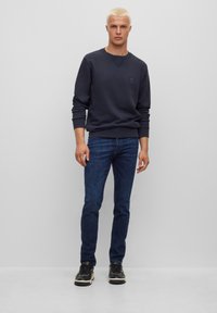 Man met kort blond haar, gekleed in een donkerblauwe sweatshirt, blauwe jeans en zwarte sneakers, staan tegen een effen lichtgrijze achtergrond.