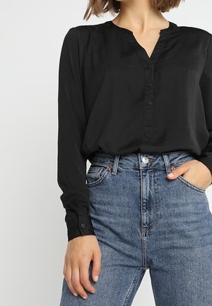 Kobieta ubrana w czarną bluzkę z długim rękawem z guzikami oraz wysokiej talii niebieskie dżinsy, z jedną ręką w kieszeni, a drugą swobodnie spuszczoną.