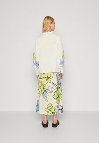 Crème blouse met lange mouwen en blauwe en groene bloemenborduursels op de mouwen, gecombineerd met een crème rok met grote bloemmotieven. Zwarte instappers.