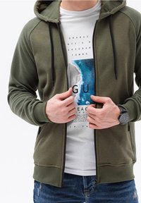 Felpa con zip verde oliva con maniche a coste, abbinata a una t-shirt bianca con grafica oceanica blu, indossata sopra jeans blu strappati.