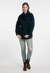 Giacca a quadri verde e blu scuro con tasche frontali, chiusure a bottoni e una cintura. Indossata sopra jeans chiari con finiture sfrangiate e stivali marroni con lacci.