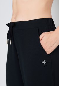 Zwarte joggingsbroek met een trekkoord in de taille, voorzakken en geborduurd logo detail aan de linkerkant. Zachte, comfortabele stof.