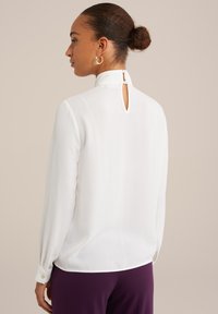 Vit blus med långa ärmar, hög halsringning och knäppning bak. Har en slät yta och en lätt glans.