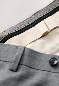 Vita di pantaloni grigi eleganti con bottone nero, fodera interna beige e bordo a quadretti visibile all'interno della vita.
