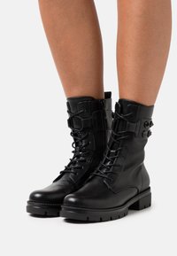 Botas de cuero negro hasta el tobillo con cordones en la parte delantera, cremalleras laterales y suelas de goma gruesa. Presentan correas decorativas y hebillas.