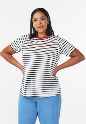 Donna con carnagione media e capelli neri che indossa una maglietta a righe bianche e nere con la scritta rossa "vibrazioni positive" e jeans blu, con le mani sui fianchi.