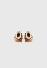 Une paire de pantoufles intérieures beiges avec un revêtement en fourrure blanche et des détails de couture en zigzag, vues de l'arrière sur un fond uni.