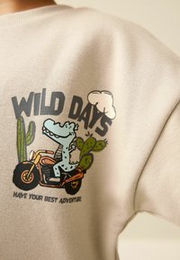 Šviesiai smėlio spalvos džemperis su karikatūrinio krokodilo grafika ant motociklo, supamas kaktusų ir debesų. Užraše parašyta „WILD DAYS“.