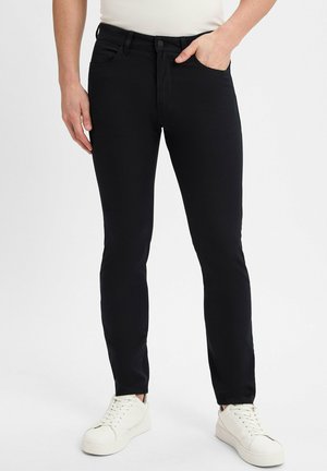 Mann, der schlanke schwarze Jeans und weiße Sneaker trägt, mit einer Hand in der Tasche vor einem schlichten Hintergrund steht.