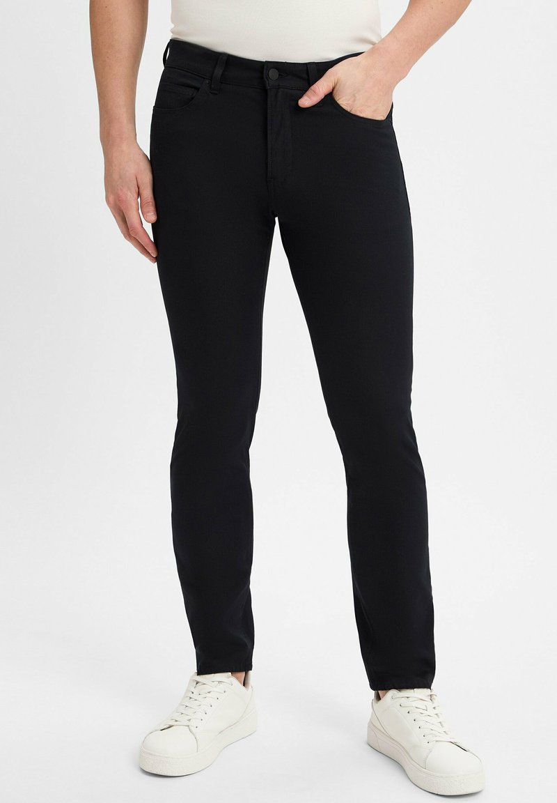 Mężczyzna w wąskich czarnych dżinsach i białych trampkach, stojący z jedną ręką w kieszeni na jednolitym tle.