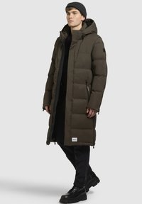 Lange olijfgroen pufferjas met capuchon, met een ritssluiting aan de voorkant, zakken aan de zijkanten en een gestructureerde stof. Draagt over een zwarte outfit en gecombineerd met zwarte laarzen.