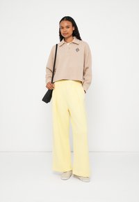 Camisola polo bege de malha com abertura de botões, combinada com calças largas amarelas de cintura alta. Mala preta de tiracolo. Ténis claros.