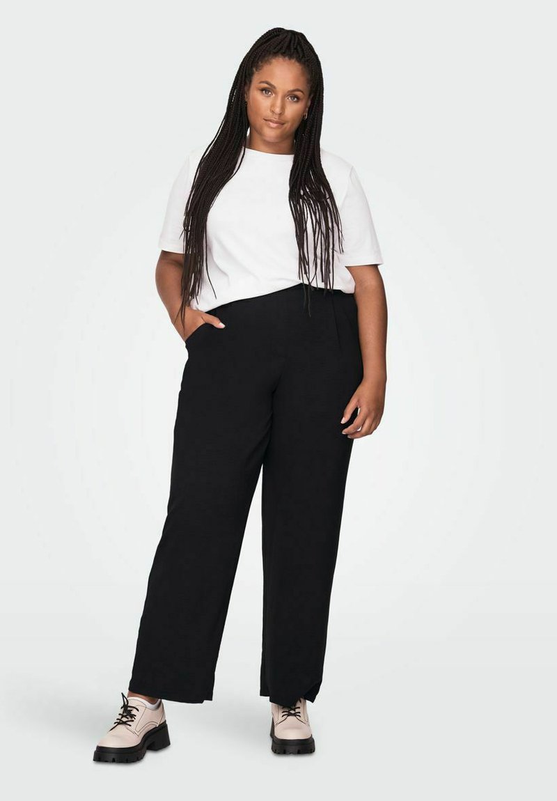 Schwarze weit geschnittene Hose mit lockerer Passform und schlichtem Design, kombiniert mit einem weißen Rundhalsausschnitt-T-Shirt und hellen Schnürschuhen.