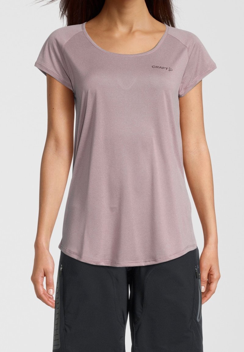 Kobieta w krótkim rękawie, w jasnofioletowej sportowej koszulce z logo "Craft" i czarnych spodniach z zamkiem, stojąca na tle białym.