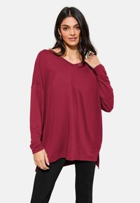 Burgundy long-sleeve top with a V-neck, relaxed fit, and side slits. Soft fabric with a smooth texture, paired with black fitted pants.