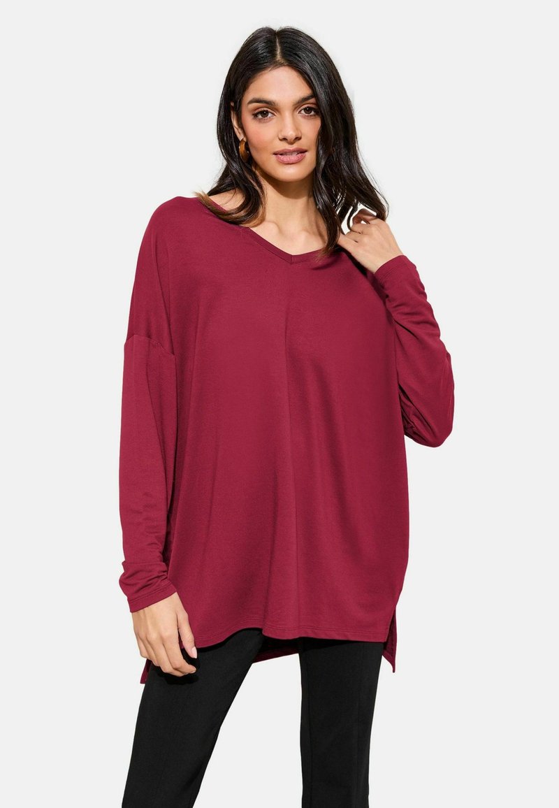 Burgundy long-sleeve top with a V-neck, relaxed fit, and side slits. Soft fabric with a smooth texture, paired with black fitted pants.