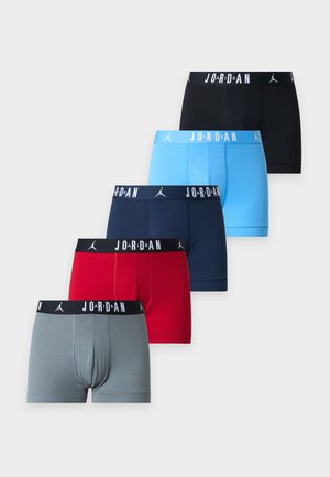 Hombre sin camiseta con cabello oscuro está descalzo vistiendo calzoncillos tipo bóxer Jordan azul claro sobre un fondo liso gris claro.