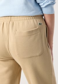Beiga sweatpants har en mjuk textur, elastisk midja och en bakficka. En liten grön Lacoste-logga syns ovanför fickan.