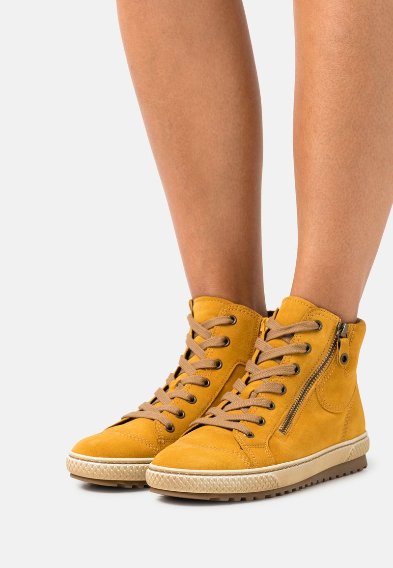 Baskets montantes jaunes avec lacets beiges et fermetures éclair latérales portées sur jambes nues sur un fond clair uni.