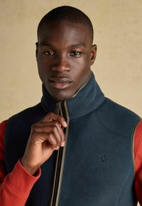 Jūras spēku vilnas vests ar augstu apkakli, ar rāvējslēdzēju priekšā un izšūtu logo; uzvilkts pāri sarkanai pitkä piedurknēm krekla. Gluda tekstūra, pieguļoša piegriezte.