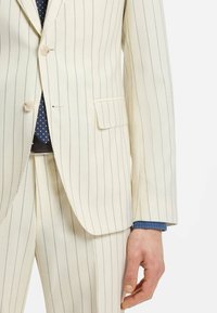 Homme portant un costume crème à fines rayures avec pantalon assorti, une cravate bleu marine à motifs et un poignet de chemise bleu visible, main détendue sur le côté.