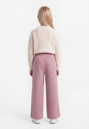 Barn med langt blondt hår står med ryggen til, iført en cremefarvet ribstrikket sweater, lyserøde vidde bukser og hvide sko på en hvid baggrund.