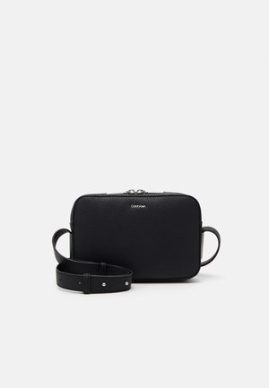 Černá texturovaná kožená kabelka Calvin Klein přes rameno s nastavitelným popruhem a stříbrným zipem na světlém pozadí.