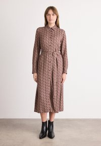 Abito con maniche lunghe e bottoni in tessuto mauve con una stampa a G. Presenta una vita con cintura e scende sotto il ginocchio. Abbinato a stivaletti neri alla caviglia.