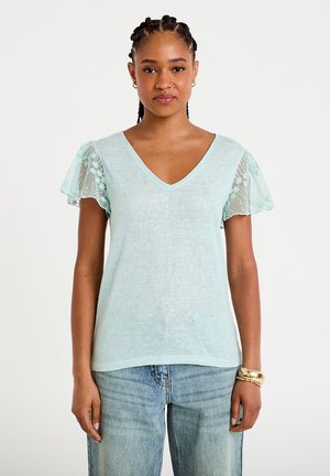 Kobieta z warkoczem na włosach ubrana w jasnoniebieską bluzkę z dekoltem w kształcie litery V, z przezroczystymi, kwiatowymi, falującymi rękawami oraz wytartymi, niebieskimi dżinsami, stoi na białym tle.