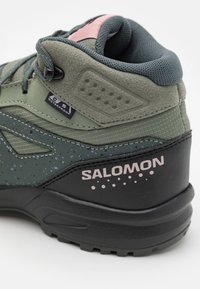 Botas de senderismo impermeables en color verde con acentos grises, superficie texturizada y detalles en rosa. Presenta el logo de "Salomon" en el talón y un diseño estampado.