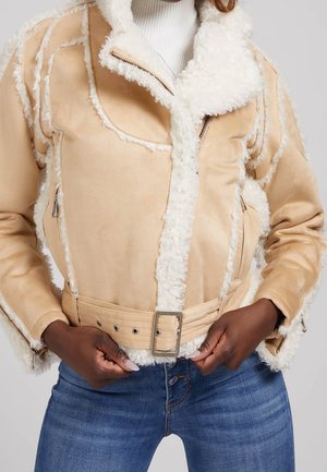 Femme attachant une veste en shearling beige avec ceinture sur un col roulé blanc, portant un jean bleu, mains positionnées près de la boucle de ceinture.