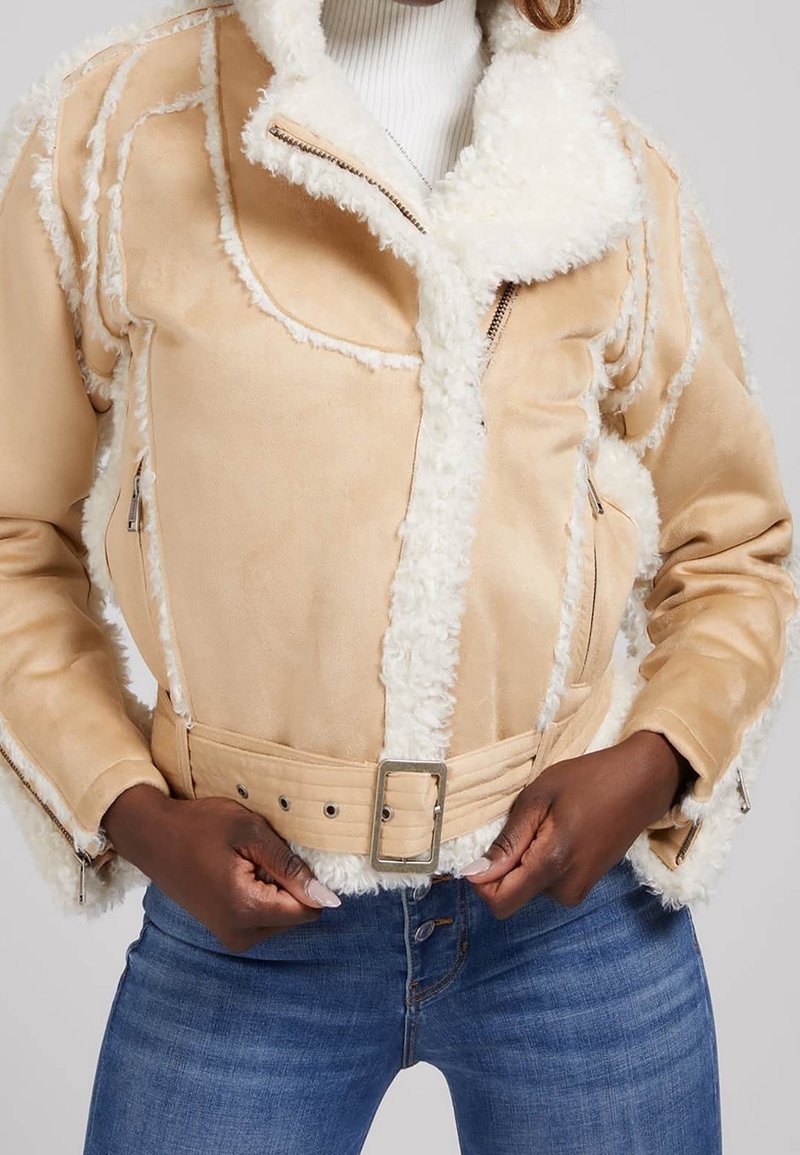 Femme attachant une veste en shearling beige avec ceinture sur un col roulé blanc, portant un jean bleu, mains positionnées près de la boucle de ceinture.