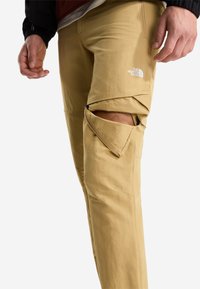 Pantaloni da trekking beige con aperture con zip sulle ginocchia, tessuto elasticizzato e logo The North Face. Presentano una trama liscia e cuciture rinforzate.