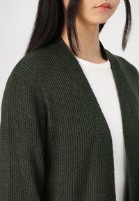 Mørkegrønn strikket cardigan med ribbestruktur og åpen frontdesign, båret over en hvit tettsittende topp. Fremtredende detaljer inkluderer en avslappet passform.