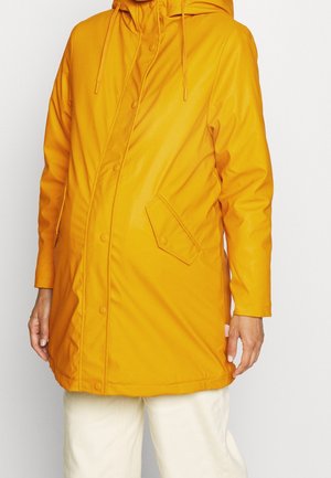 Impermeabile giallo con cappuccio, chiusura a bottoni automatici e tasche con patta obliqua, indossato sopra pantaloni bianchi, visto dalla metà coscia fino al collo.