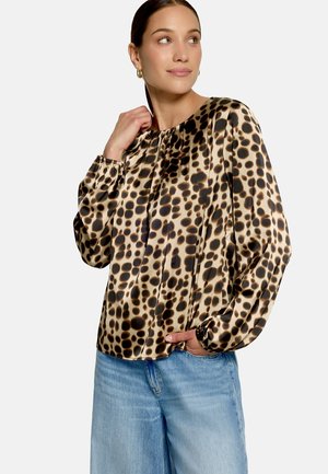 Femme portant un chemisier beige à manches longues avec des taches irrégulières noires et marron, associé à un jean en denim bleu clair.