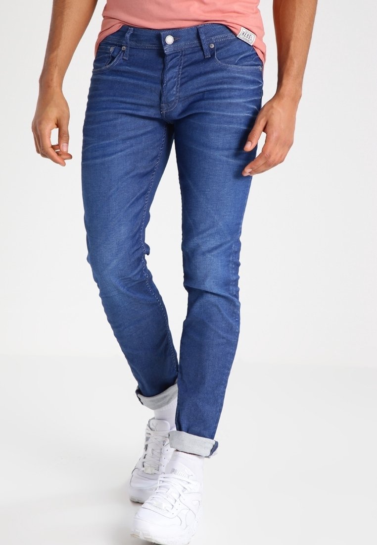 Man som bär uppvikta slim-fit blå jeans, vita sneakers och vita strumpor, står med avslappnade händer vid sidorna.