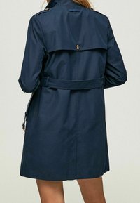 Mörkblå trenchcoat i ett slätt tyg, med bälte i midjan, axelklaffar och en knäppt bakklaff för ventilation.