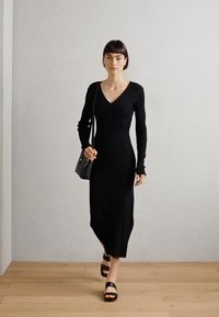 Robe longue noire en maille côtelée avec un décolleté en V, des manches longues et des détails de boutons. Associée à des sandales noires et un petit sac à main.