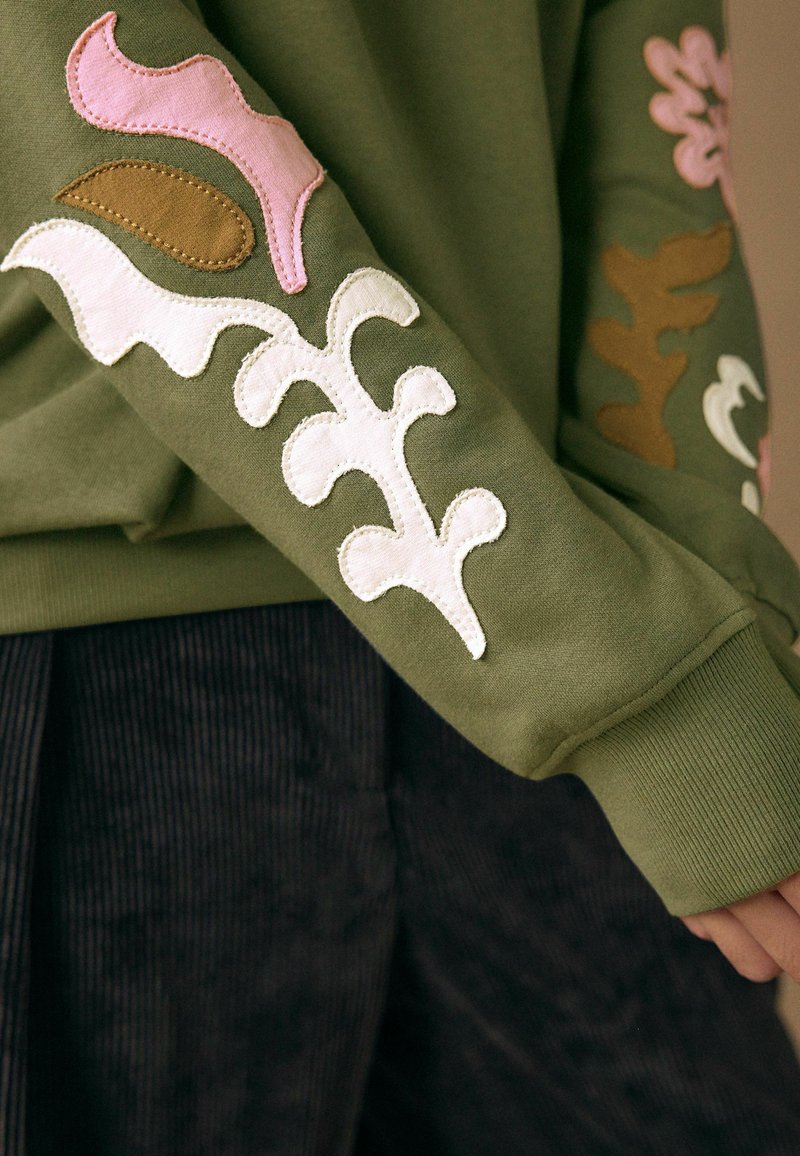 Sweat-shirt vert olive avec broderie florale blanche, rose et marron sur les manches. Texture lisse avec poignets côtelés.