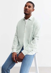 Camisa a rayas verde claro con un ajuste relajado, bolsillo en el pecho, y cierres de botones, combinada con jeans de mezclilla azules.